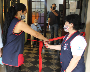 Duas mulheres cortando uma fita vermelha durante uma cerimônia de inauguração, com pessoas observando ao fundo